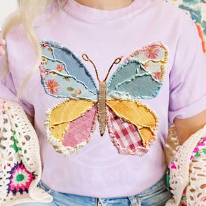 May include: A light purple t-shirt featuring a large, colorful butterfly applique. The butterfly has patchwork wings in shades of blue, yellow, pink, and red, with floral and gingham patterns. The antennae are brown. The shirt is a casual style.