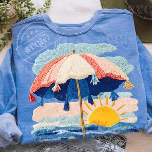 May include: A light blue sweatshirt featuring a colorful beach scene. The design includes a striped beach umbrella, a setting sun, and ocean waves. The umbrella has tassels and is made of various colors, including pink, blue, and cream.