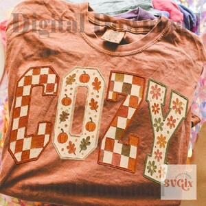 May include: A terracotta-colored t-shirt featuring the word COZY printed in large block letters. The C and Z have a brown and white checkered pattern, the O contains orange pumpkins and leaves, and the Y features orange and green flowers.