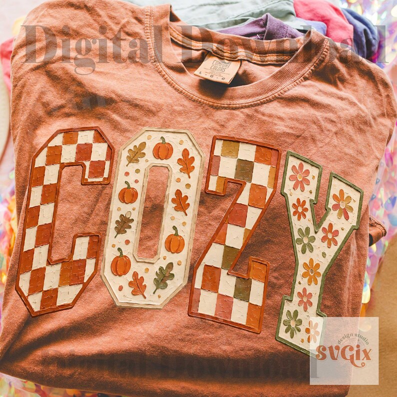 May include: A terracotta-colored t-shirt featuring the word COZY printed in large block letters. The C and Z have a brown and white checkered pattern, the O contains orange pumpkins and leaves, and the Y features orange and green flowers.