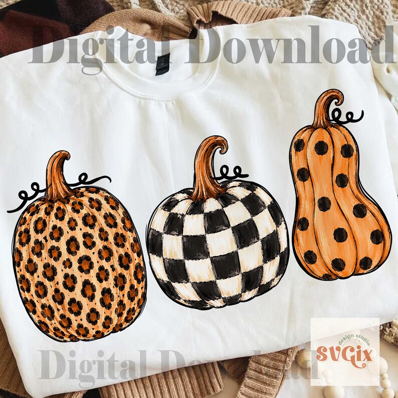 Puede incluir: Sudadera blanca con tres dise&ntilde;os de calabazas. Una calabaza tiene un estampado de leopardo, otra un patr&oacute;n de tablero de ajedrez en blanco y negro, y la tercera es naranja con lunares negros. Las calabazas tienen tallos marrones y zarcillos negros rizados.