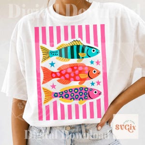 May include: White t-shirt featuring a colorful graphic of three stylized fish. The fish are in shades of blue, red, and pink, with yellow accents and star details. The design is set against a pink striped background.