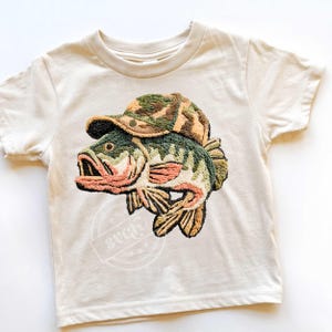 May include: A cream-coloured t-shirt with an embroidered design of a fish wearing a camouflage hat. The fish is detailed with green, brown, and peach colours. The shirt has a round neck and short sleeves, suitable for children.