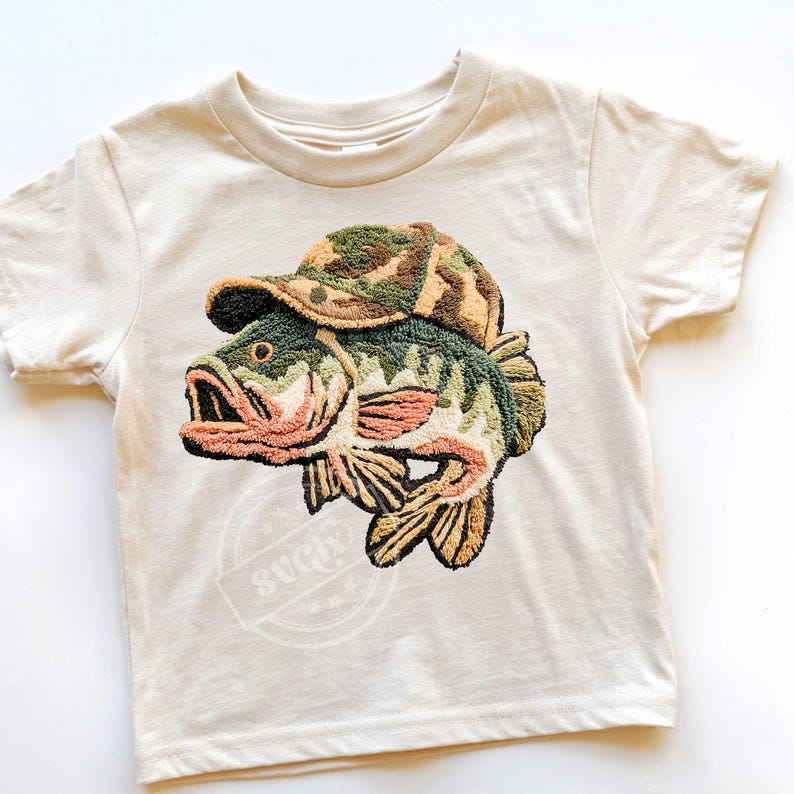 May include: A cream-coloured t-shirt with an embroidered design of a fish wearing a camouflage hat. The fish is detailed with green, brown, and peach colours. The shirt has a round neck and short sleeves, suitable for children.