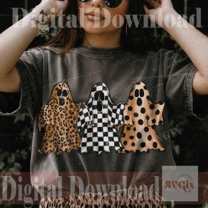 May include: A charcoal gray short-sleeve t-shirt featuring a graphic of three ghosts. The left ghost has a brown leopard print, the center ghost has a black and white checkerboard pattern, and the right ghost has a brown polka dot pattern.