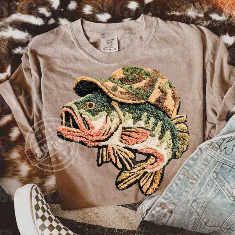 May include: A tan t-shirt with an embroidered design of a fish wearing a camouflage hat. The fish is green, orange, and yellow. The shirt has a crew neck and short sleeves. The tag on the shirt reads "Comfort Colors".