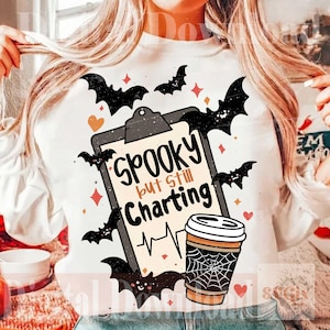 May include: A white sweatshirt featuring a graphic of a clipboard with the text "Spooky but Still Charting" above a heartbeat line. The design includes a coffee cup with a spiderweb pattern, black bats, and small orange and red hearts and stars.