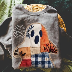 May include: Gray sweatshirt with a patchwork ghost design. The ghost features a white face with three dark blue eye cutouts, orange accents, and a black spiderweb. The word "ORIGINAL" is printed on the upper left. The word "S" is printed on the lower right.