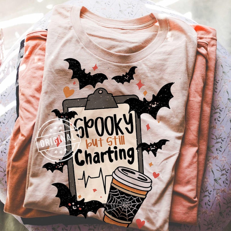 May include: A stack of folded long-sleeved shirts. The top shirt is light beige with a Halloween-themed graphic that includes bats, hearts, and the text "Spooky but still Charting." The graphic also features a coffee cup with a spiderweb design.