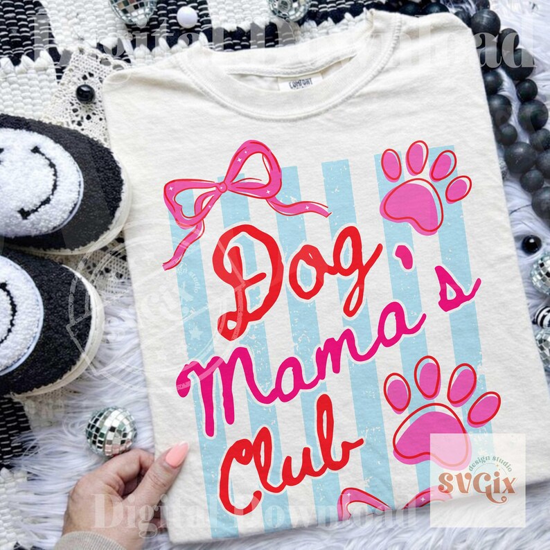 May include: Cream-colored t-shirt with the text "Dog Mama's Club" in red and pink. The design includes a pink bow, paw prints, and light blue vertical stripes. The shirt is a casual wear item.