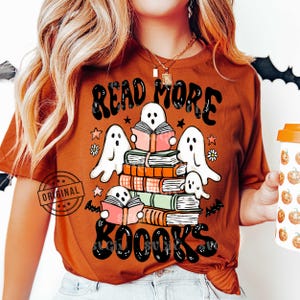 Peut inclure: T-shirt orange brûlé avec l'inscription "READ MORE BOOKS" et des illustrations de fantômes lisant des livres. Le motif comprend des étoiles, des fleurs et des chauves-souris. Le t-shirt a un col rond et des manches courtes.