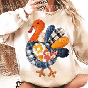 May include: A cream-colored sweatshirt featuring a colorful turkey applique. The turkey has an orange head and beak, a blue plaid body, and a patchwork of patterned feathers in orange, black, and white. Brown turkey feet complete the design.