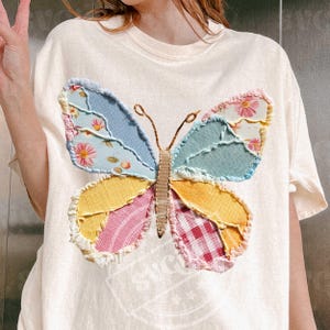 May include: Cream-colored t-shirt featuring a large, colorful butterfly applique. The butterfly wings are patchwork, with sections of blue, yellow, pink, and floral patterns. The antennae are thin, brown lines. The shirt has a round neckline and short sleeves.