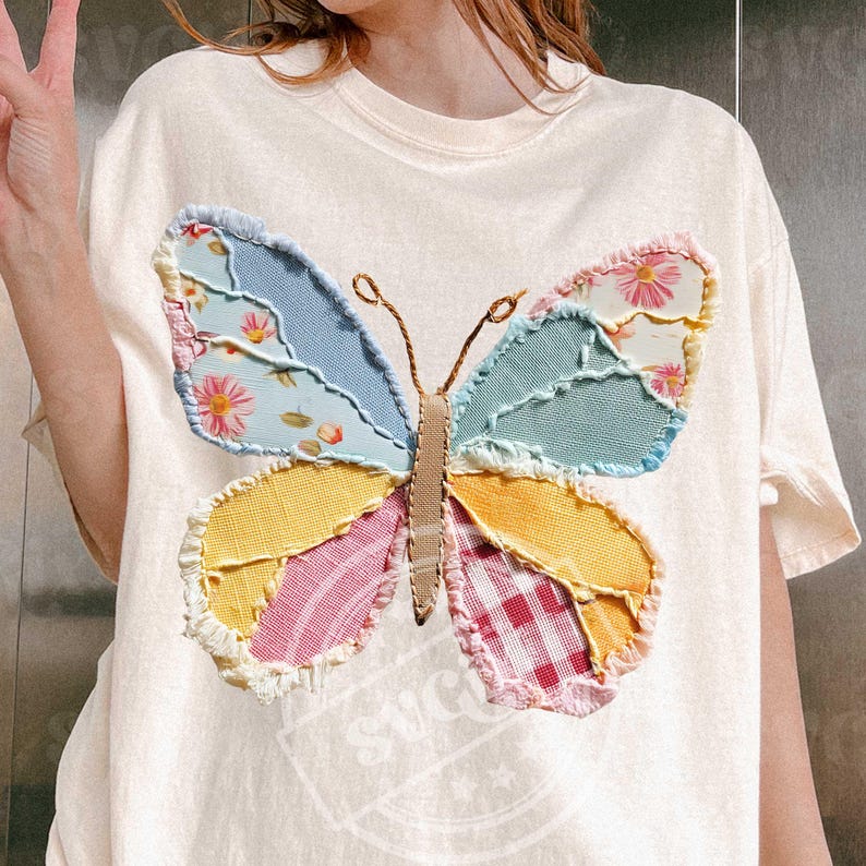 May include: Cream-colored t-shirt featuring a large, colorful butterfly applique. The butterfly wings are patchwork, with sections of blue, yellow, pink, and floral patterns. The antennae are thin, brown lines. The shirt has a round neckline and short sleeves.