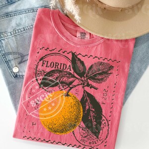 May include: A coral t-shirt featuring a graphic of an orange and the word "FLORIDA" in a stamp design. The shirt is folded on blue jeans, with a necklace and a tan hat visible.