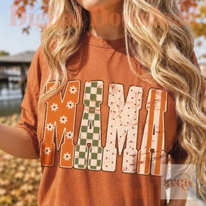 May include: A rust-colored t-shirt featuring the word MAMA in large block letters. The first letter has a white daisy pattern, the second has a green and white checkerboard print, and the last two have a distressed light pink polka dot design.