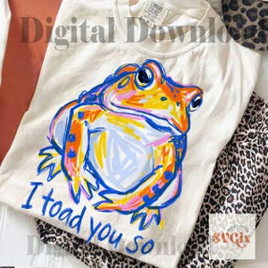 May include: Cream-colored t-shirt with a colorful frog illustration. The frog is depicted in orange, blue, yellow, and pink. The text "I toad you so" is written in blue below the frog. The shirt is folded and placed on a surface with leopard print fabric.