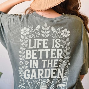 Puede incluir: Camiseta verde salvia con la frase "LIFE IS BETTER IN THE GARDEN" en letras blancas. El dise&ntilde;o incluye ilustraciones florales y de p&aacute;jaros, con una peque&ntilde;a silla y un cubo de flores en la parte inferior.