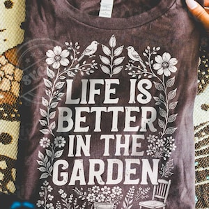 Puede incluir: Camiseta marr&oacute;n oscuro con el texto blanco "LIFE IS BETTER IN THE GARDEN". El dise&ntilde;o incluye elementos florales y de hojas, dos p&aacute;jaros y una peque&ntilde;a silla. La camiseta es de cuello redondo.