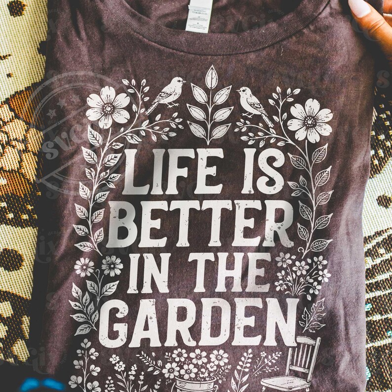 Puede incluir: Camiseta marr&oacute;n oscuro con el texto blanco "LIFE IS BETTER IN THE GARDEN". El dise&ntilde;o incluye elementos florales y de hojas, dos p&aacute;jaros y una peque&ntilde;a silla. La camiseta es de cuello redondo.