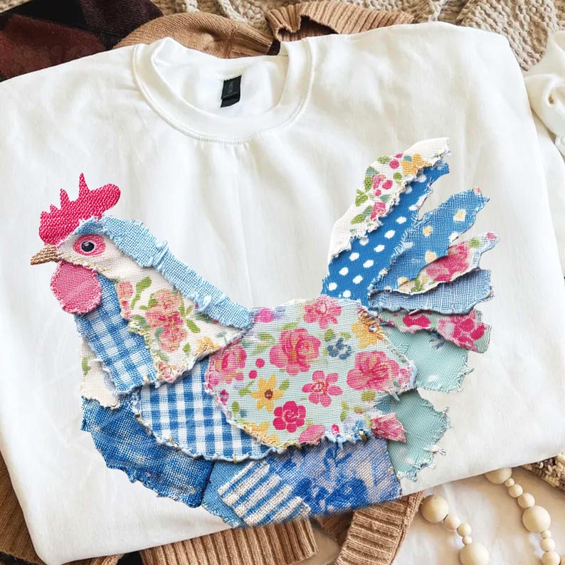 May include: White crewneck sweatshirt with a colorful patchwork rooster design. The rooster features various fabric patterns, including floral, polka dot, and gingham, in shades of blue, pink, and white. The rooster's comb is bright pink.