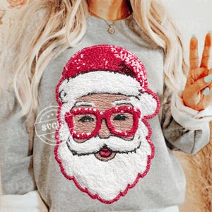 Könnte beinhalten: Ein graues Sweatshirt mit einem paillettenbesetzten Weihnachtsmann-Gesicht. Das Santa-Design umfasst einen roten Hut und eine Brille, einen weißen Bart und einen lächelnden Mund. Dies ist ein festliches Kleidungsstück für die Feiertage.