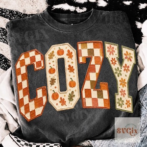 May include: A charcoal gray crewneck sweatshirt featuring the word COZY in large block letters. The C is orange and white checkered, the O features orange pumpkins and leaves, the Z is orange and brown checkered, and the Y has floral patterns.