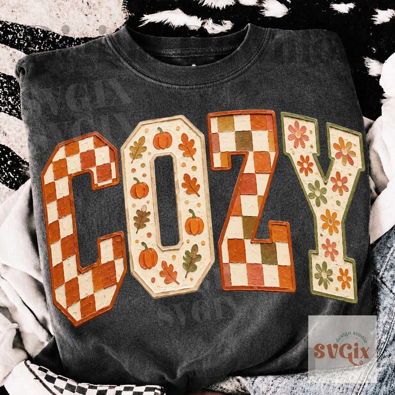 May include: A charcoal gray crewneck sweatshirt featuring the word COZY in large block letters. The C is orange and white checkered, the O features orange pumpkins and leaves, the Z is orange and brown checkered, and the Y has floral patterns.