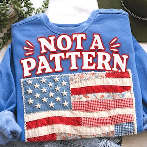 Pu&ograve; includere: Una felpa blu chiaro con la scritta "NOT A PATTERN" in bianco e rosso. La parte anteriore presenta una bandiera americana patchwork con stelle bianche su campo blu e strisce rosse e floreali.