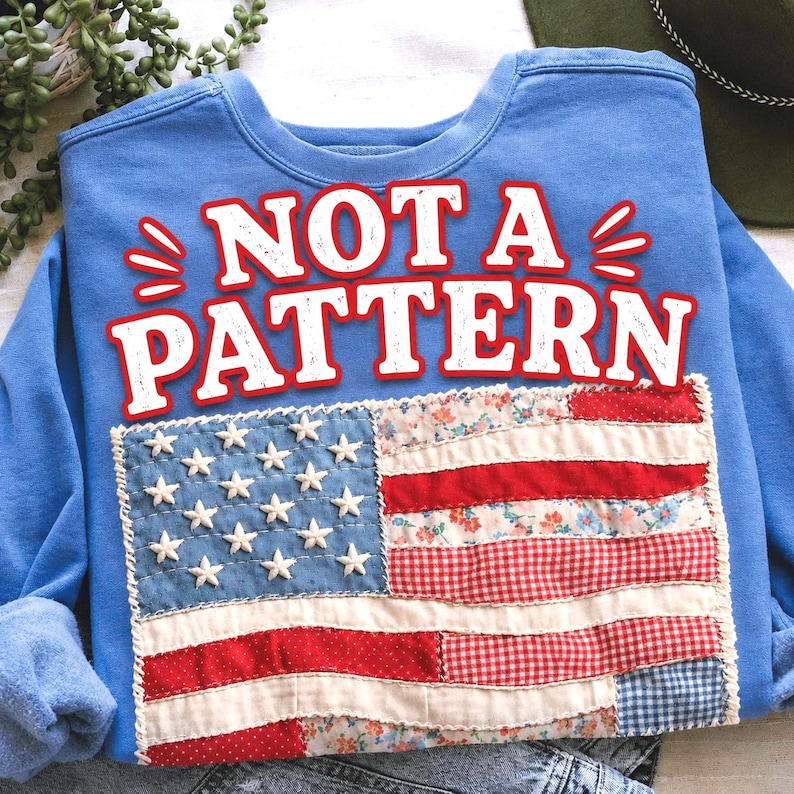 Pu&ograve; includere: Una felpa blu chiaro con la scritta "NOT A PATTERN" in bianco e rosso. La parte anteriore presenta una bandiera americana patchwork con stelle bianche su campo blu e strisce rosse e floreali.