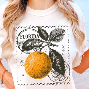 May include: Off-white t-shirt with a retro graphic of an orange, leaves, and the word "FLORIDA" in a circular stamp. The design is in black and orange, giving it a vintage feel.