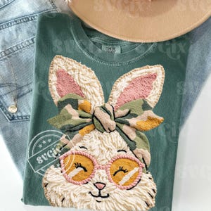May include: A green t-shirt with a detailed bunny design wearing sunglasses and a camouflage bow. The bunny has pink inner ears and a fluffy, textured appearance. The shirt is folded with blue jeans and a hat in the background.