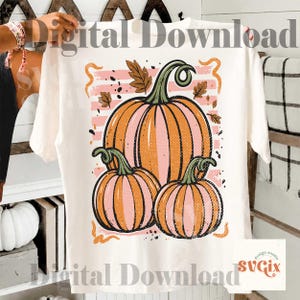 May include: Cream-colored t-shirt featuring a fall-themed graphic. The design includes three pumpkins in shades of orange and pink, with green stems and leaves. The words "Digital Download" are printed on the shirt.