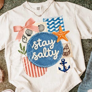 Peut inclure: T-shirt blanc orn&eacute; de motifs marins : coquillages, vagues et &eacute;toile de mer orange. Un &eacute;cusson bleu avec l'inscription "stay salty" en blanc est au centre. Un n&oelig;ud rose et un collier argent&eacute; compl&egrave;tent le design.