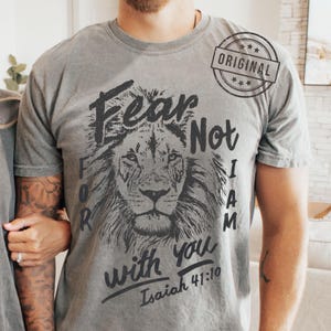 Puede incluir: Camiseta gris con una ilustraci&oacute;n de le&oacute;n y el texto "Fear Not For I Am With You" y el vers&iacute;culo Isa&iacute;as 41:10. La camiseta tambi&eacute;n tiene un sello "ORIGINAL".