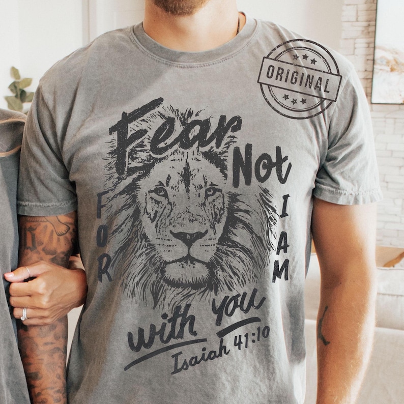 Puede incluir: Camiseta gris con una ilustraci&oacute;n de le&oacute;n y el texto "Fear Not For I Am With You" y el vers&iacute;culo Isa&iacute;as 41:10. La camiseta tambi&eacute;n tiene un sello "ORIGINAL".