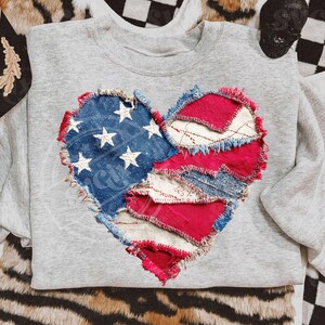 May include: A grey sweatshirt with a heart-shaped patch made from denim and red fabric. The denim section features white stars, similar to the American flag. The edges of the heart are frayed.