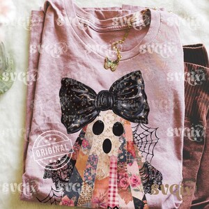 May include: A mauve-colored t-shirt featuring a ghost design with a large black bow and patchwork body. The ghost has three black eye holes and is decorated with floral and patterned fabrics. A gold butterfly necklace is also visible.