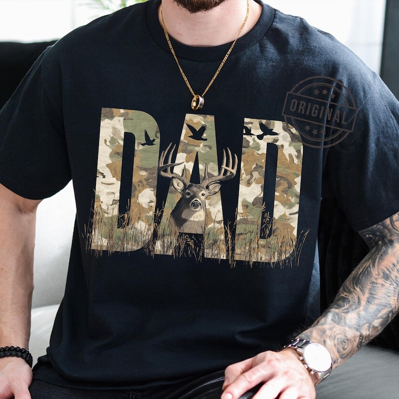 May include: A black t-shirt with a camouflage pattern featuring the word "DAD" with a deer in the middle of the word. The deer is brown with large antlers.