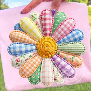 Puede incluir: Una colorida flor de tela hecha a mano con un centro bordado amarillo y pétalos multicolores. Los pétalos presentan varios patrones, incluyendo cuadros, lunares y estampados florales, con un borde cosido. La flor está sobre un fondo rosa.