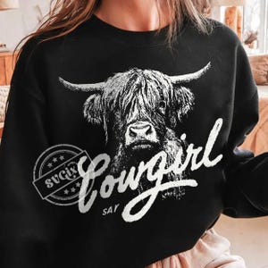 May include: Black sweatshirt featuring a white graphic of a Highland cow with the word "Cowgirl" in a script font. The design includes a circular stamp with the text "SVGix" and stars.