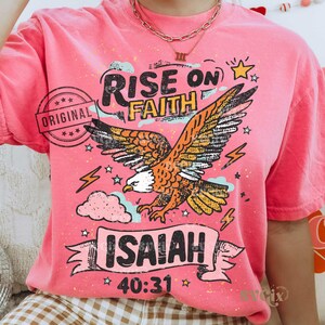 Puede incluir: Camiseta rosa con un gr&aacute;fico de un &aacute;guila con las alas extendidas, las palabras "RISE ON FAITH" e "ISAIAH 40:31". El &aacute;guila es naranja, marr&oacute;n y blanca. La camiseta tiene un aspecto vintage y desgastado.