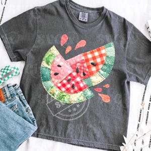 Puede incluir: Camiseta gris oscuro con un diseño de rodaja de sandía colorida. La sandía está hecha de tela de patchwork con patrones rojos, rosas, verdes y blancos. Semillas negras y acentos de gotas rojas completan el diseño. La camiseta se muestra con vaqueros azules.