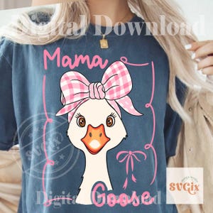 Pode incluir: Camiseta azul com um desenho de ganso de desenho animado usando um la&ccedil;o e uma faixa xadrez rosa e branca. As palavras "Mama Goosed" est&atilde;o escritas em rosa. O desenho &eacute; emoldurado por linhas rosa e uma pequena fita.