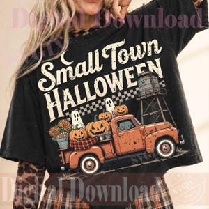 May include: A black t-shirt featuring a graphic print with the text Small Town Halloween. The design includes an orange pickup truck, pumpkins, ghosts, a water tower, and a checkered pattern. The shirt is worn over a leopard print long-sleeve top.