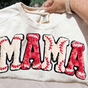 May include: Cream-colored sweatshirt with the word "MAMA" in large, textured letters. The letters are designed to resemble baseballs, with red stitching and a black outline. A stylish and comfortable top.