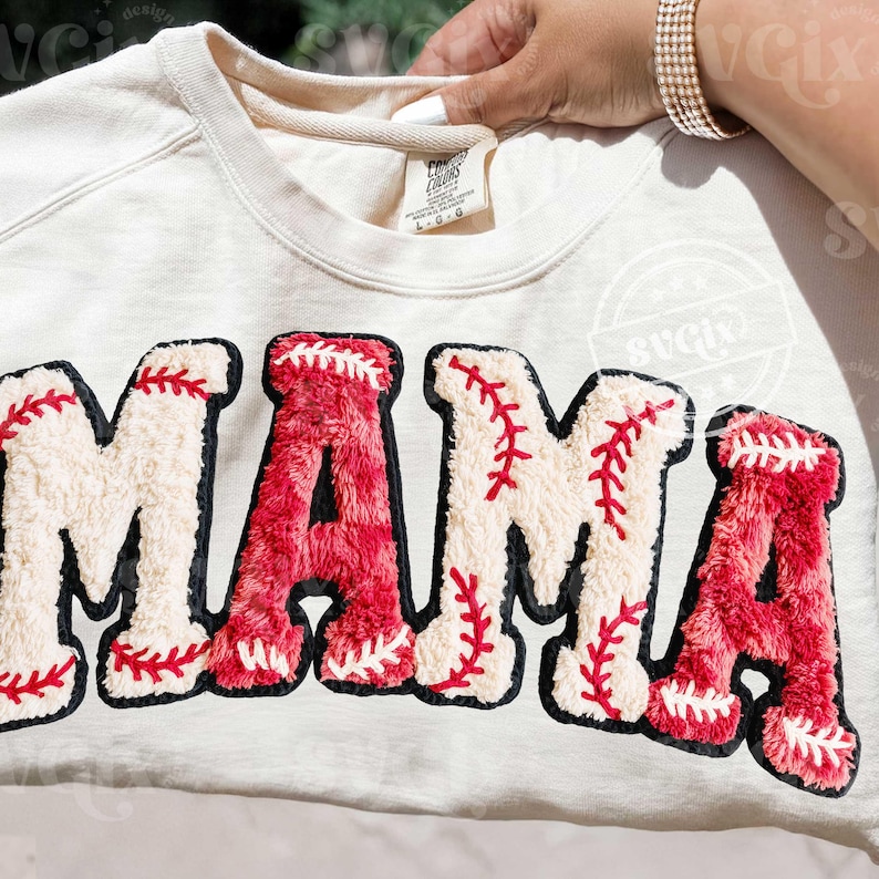 May include: Cream-colored sweatshirt with the word "MAMA" in large, textured letters. The letters are designed to resemble baseballs, with red stitching and a black outline. A stylish and comfortable top.