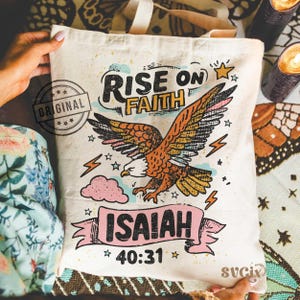 Puede incluir: Bolso tote color crema con un gr&aacute;fico de un &aacute;guila y las palabras "RISE ON FAITH" e "ISAIAH 40:31". El &aacute;guila est&aacute; representada con plumas naranjas, marrones y amarillas. La bolsa tiene un sello que dice "ORIGINAL".