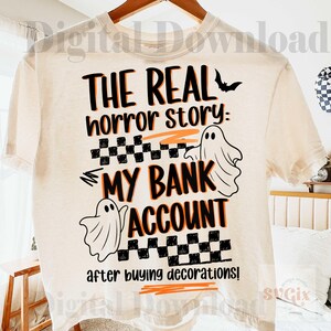 Pode incluir: Camiseta creme com um design tem&aacute;tico de Halloween. O design apresenta o texto "THE REAL horror story: MY BANK ACCOUNT after buying decorations!" em preto e laranja, com ilustra&ccedil;&otilde;es de fantasmas e um morcego.