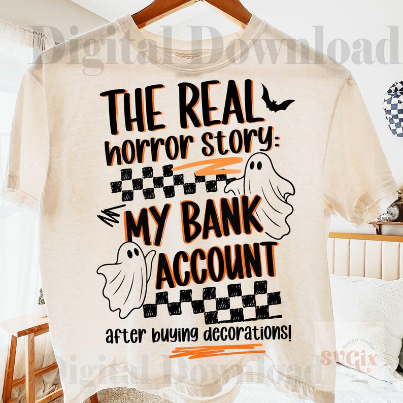 Pode incluir: Camiseta creme com um design tem&aacute;tico de Halloween. O design apresenta o texto "THE REAL horror story: MY BANK ACCOUNT after buying decorations!" em preto e laranja, com ilustra&ccedil;&otilde;es de fantasmas e um morcego.
